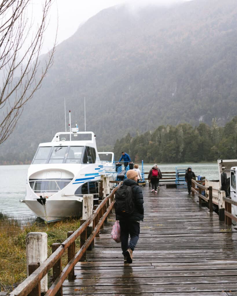 Cruce Andino: The Scenic Route From Chile to Argentina 25 Brayden walks along a wet wooden dock in Puerto Frías, carrying a backpack and a floral bag, heading toward a white and blue boat moored at the edge of the lake