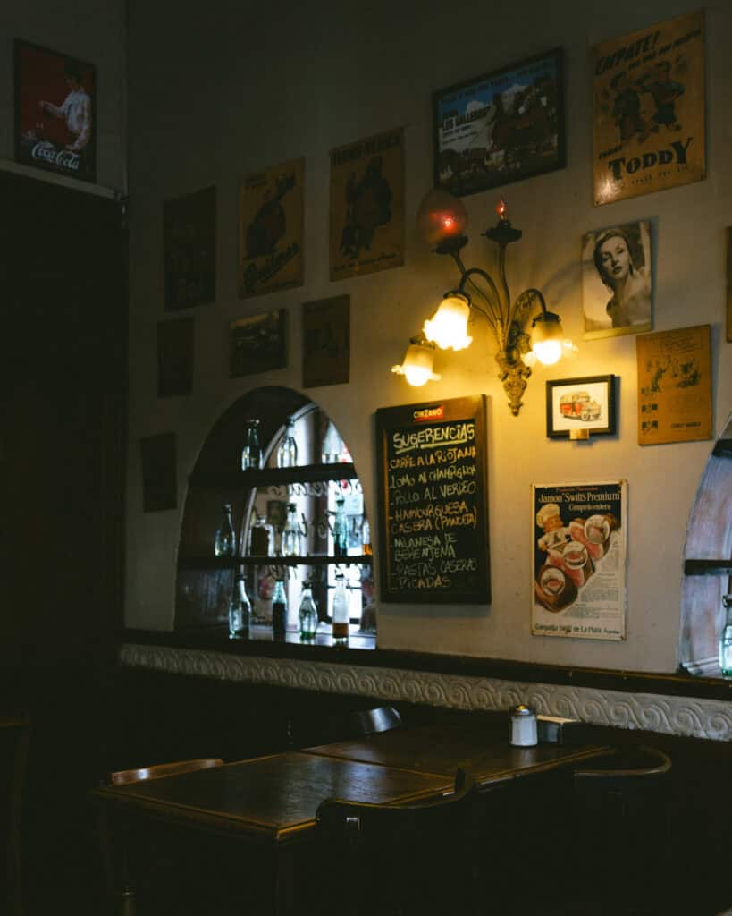 Buenos Aires Food Tour: Sherpa Food Tours in San Telmo 7 A warmly lit corner of Bar El Federal with empty wooden tables and chairs beneath retro Argentine posters and a chalkboard menu listing daily specials like pollo al verdeo and milanesa de berenjena.