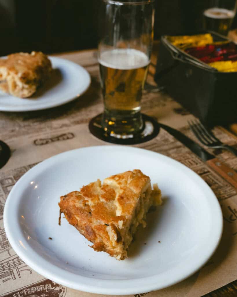 Buenos Aires Food Tour: Sherpa Food Tours in San Telmo 9 A slice of savory pie, possibly tortilla or Spanish-style potato omelette, is served on a white plate with a glass of beer in the background at Bar El Federal. The placemat features the café’s illustrated branding.