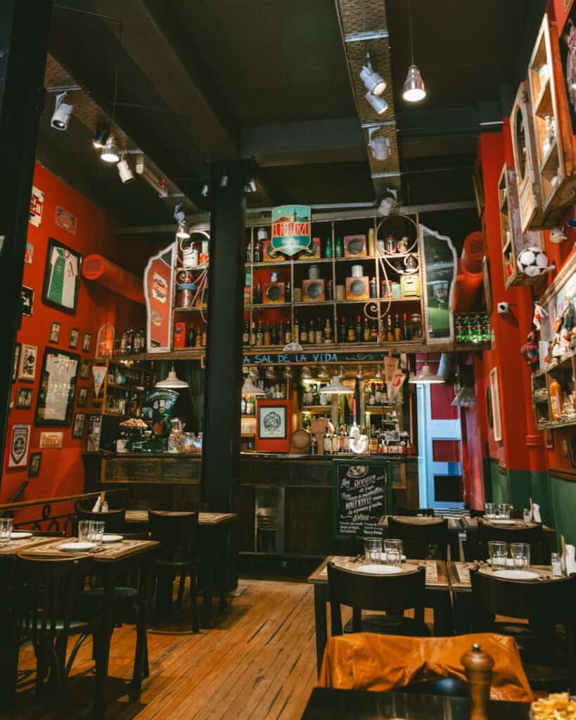 Buenos Aires Food Tour: Sherpa Food Tours in San Telmo 20 The interior of a vibrant Buenos Aires restaurant featuring red and green walls decorated with soccer memorabilia, vintage bottles, and signs like “La Sal de la Vida.” Set tables line the wooden floor beneath hanging lights.