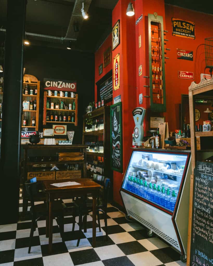 Buenos Aires Food Tour: Sherpa Food Tours in San Telmo 5 A cozy deli corner with black-and-white checkered flooring, red walls decorated with vintage beverage ads, and a refrigerated case filled with cheeses and soda siphon bottles. Wooden shelves behind the counter are stocked with wine and liquor.