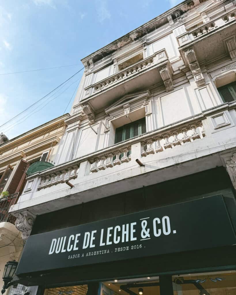 Buenos Aires Food Tour: Sherpa Food Tours in San Telmo 16 Upward view of the storefront for Dulce de Leche & Co., featuring a modern black awning and white letters reading “Sabor a Argentina. Desde 2016,” beneath a weathered, neoclassical-style building façade.