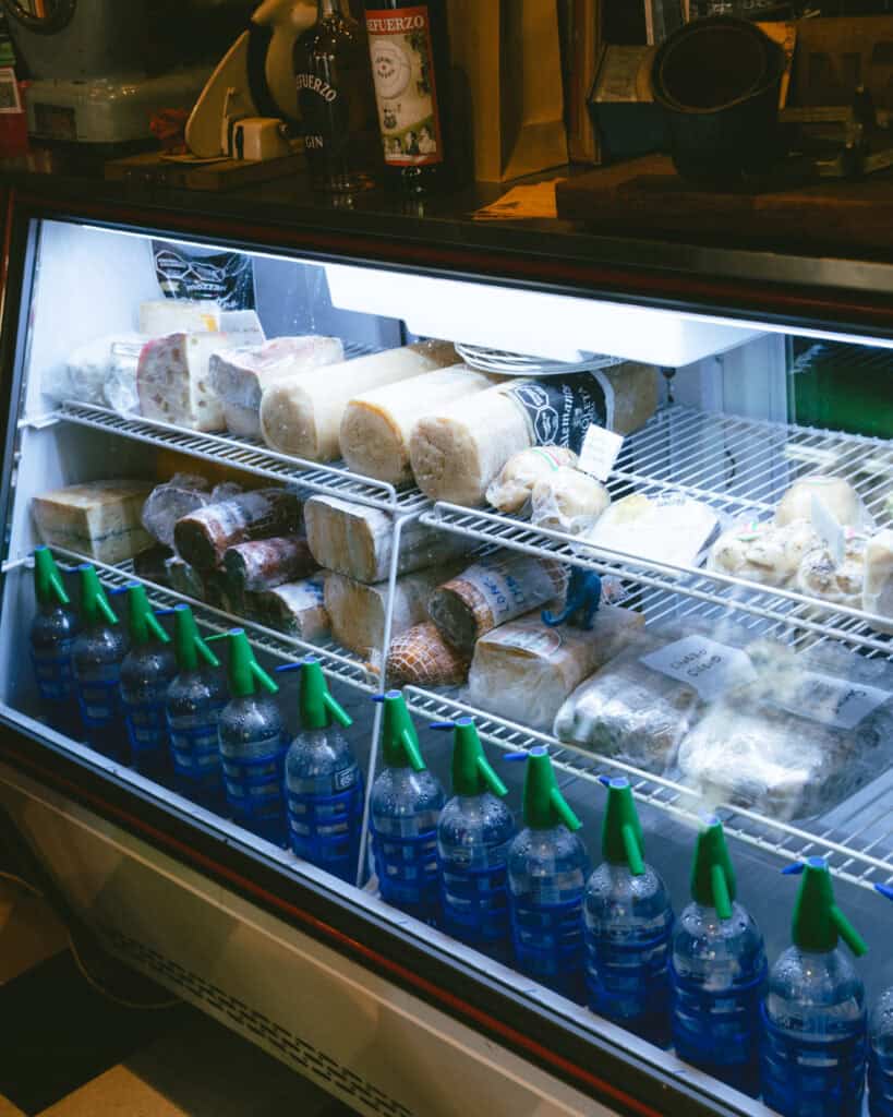 Buenos Aires Food Tour: Sherpa Food Tours in San Telmo 14 Close-up of a deli fridge stocked with various blocks and rounds of cured cheeses and cold cuts, labeled with handwritten tags. Below, rows of classic Argentine soda siphon bottles line the bottom shelf.