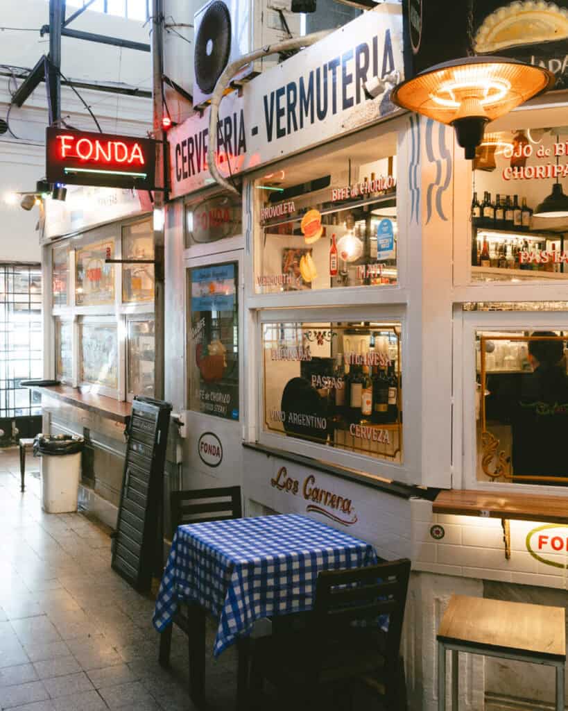 Buenos Aires Food Tour: Sherpa Food Tours in San Telmo 10 Small corner fonda inside Mercado de San Telmo with a blue checkered tablecloth on a two-seat table outside a shop labeled “Cervecería - Vermutería” serving provoleta, bife de chorizo, empanadas, and vermouth.