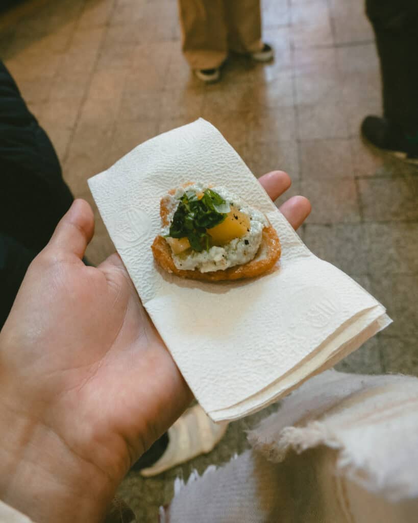Buenos Aires Food Tour: Sherpa Food Tours in San Telmo 11 Hand holding a small appetizer on a napkin, featuring a crispy toast topped with a creamy cheese spread, diced yellow tomatoes, and chopped herbs.