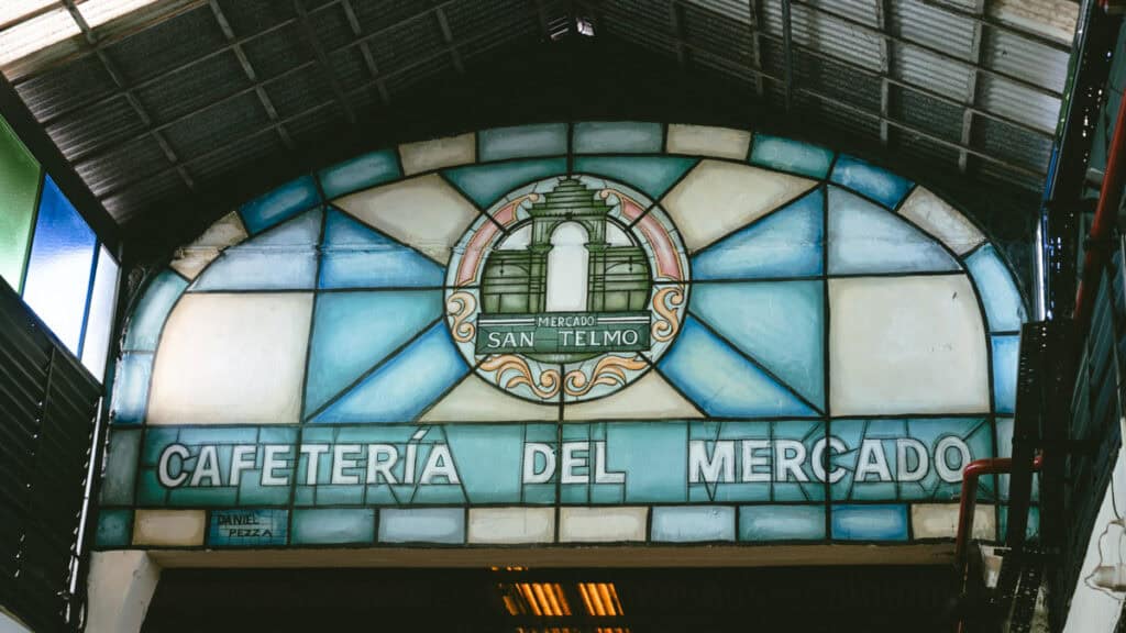 Buenos Aires Food Tour: Sherpa Food Tours in San Telmo 6 Stained glass window arch at Mercado de San Telmo reading “Cafetería del Mercado” with a painted emblem of the market at its center, framed by iron and colored panes.