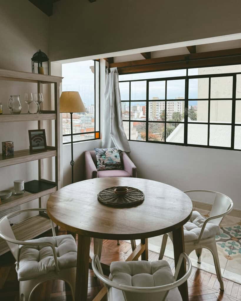 7 - 10 Day Salta Road Trip: The Best of Northwest Argentina 20 Cozy apartment dining nook in Salta City Argentina with a round wooden table, white cushioned chairs, and a corner chair by large windows offering a city view through black-framed glass panes.