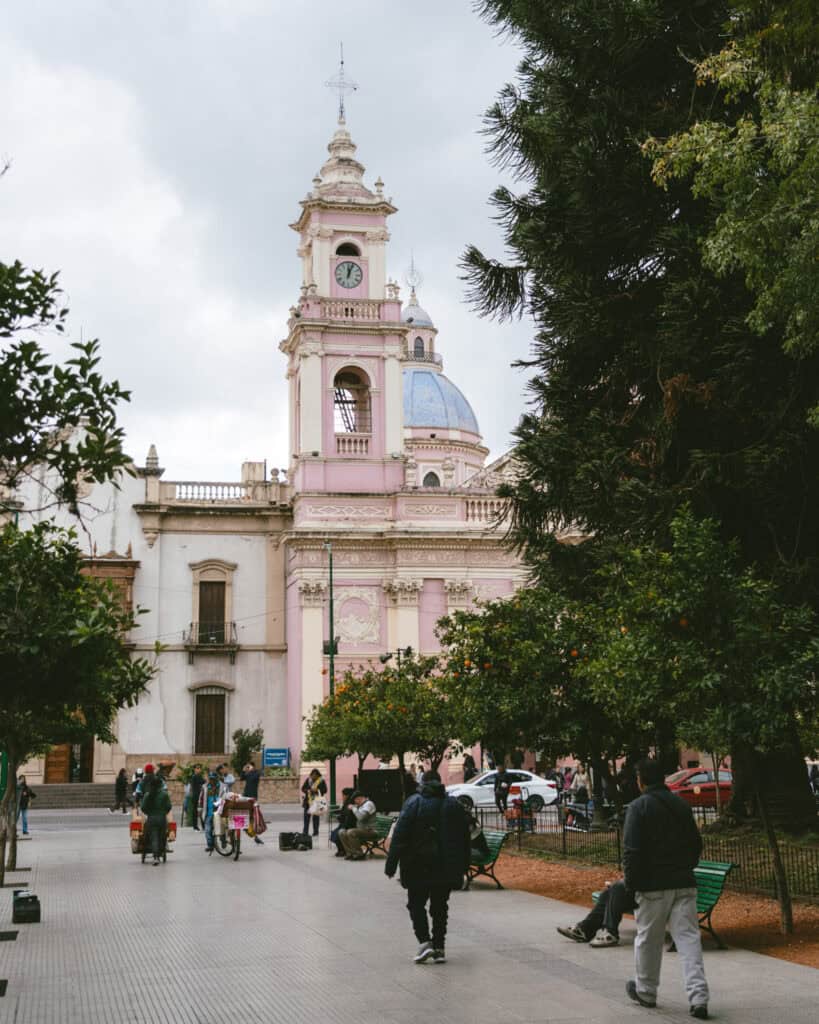7 - 10 Day Salta Road Trip: The Best of Northwest Argentina 10 Plaza in Salta City Argentina in front of a pastel pink colonial-style church with a clock tower and blue dome, surrounded by people walking, sitting on benches, and a few market carts under orange trees.