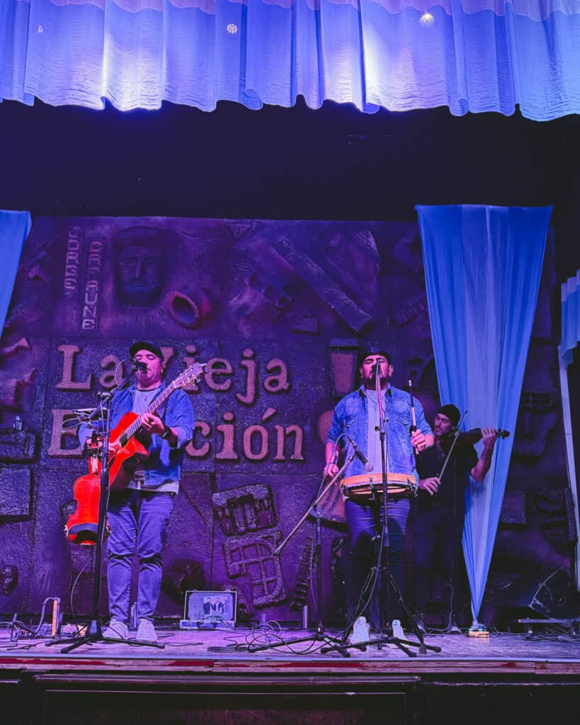 7 - 10 Day Salta Road Trip: The Best of Northwest Argentina 9 Three musicians performing at La Vieja Estación in Salta, with one playing a guitar, one on bombo drum, and another on violin beneath a purple-lit stage.