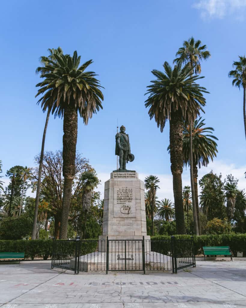 7 - 10 Day Salta Road Trip: The Best of Northwest Argentina 11 Monument to General Martín Miguel de Güemes standing tall on a stone pedestal surrounded by palm trees and green benches in Salta’s Plaza Güemes.