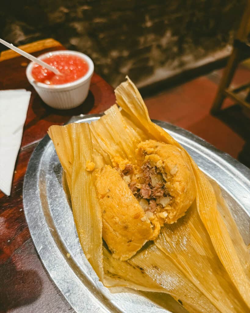 7 - 10 Day Salta Road Trip: The Best of Northwest Argentina 19 Traditional Argentine tamale served in a corn husk on a silver platter, partially opened to reveal a savory filling of meat and vegetables, with red salsa on the side.