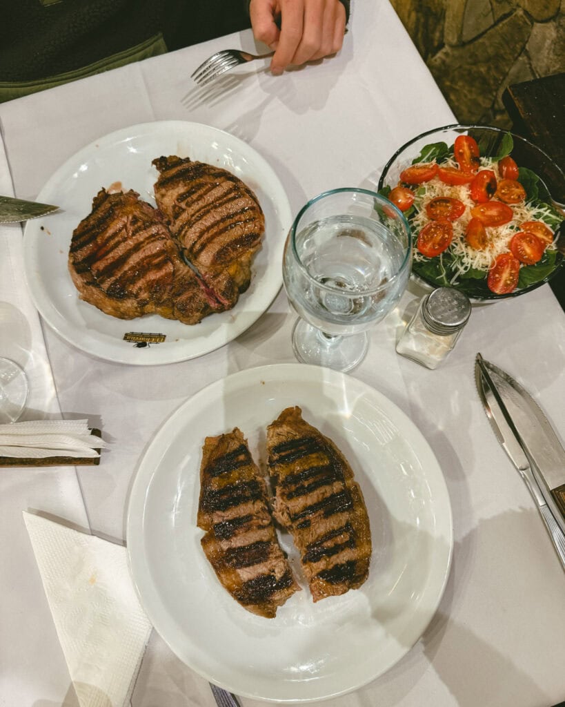 7 - 10 Day Salta Road Trip: The Best of Northwest Argentina 14 Overhead view of a steak dinner with two grilled steaks on white plates, a fresh salad with cherry tomatoes, a glass of water, and utensils on a white tablecloth.