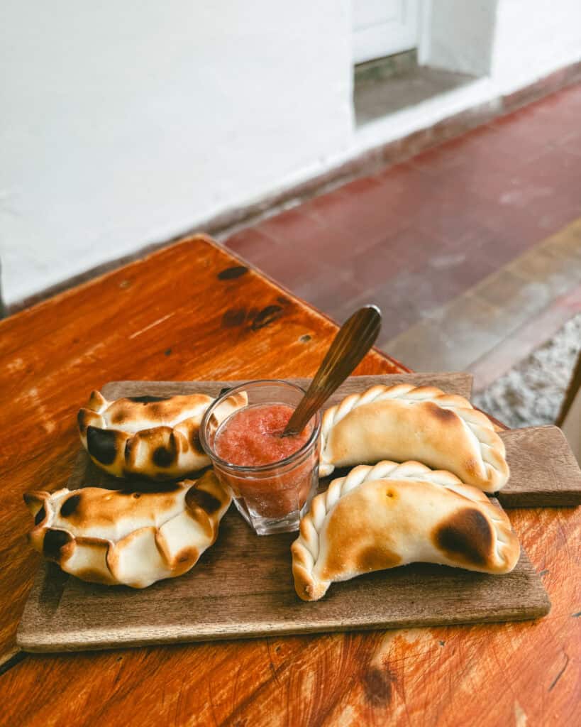 7 - 10 Day Salta Road Trip: The Best of Northwest Argentina 16 Four golden-baked empanadas served on a wooden board with a glass of red salsa, set on a rustic wooden table near a white stucco wall.