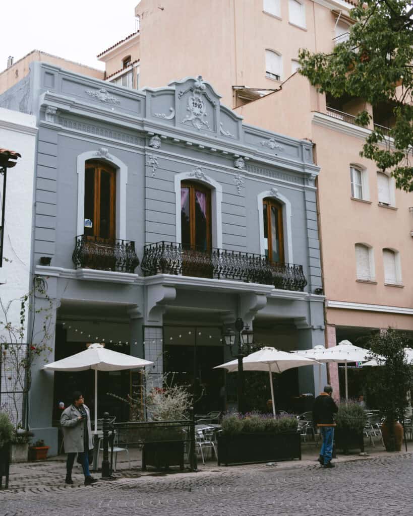 7 - 10 Day Salta Road Trip: The Best of Northwest Argentina 13 Two-story neoclassical building in Salta painted soft gray with ornate trim and “1910” marked at the top, featuring French balconies and an outdoor café setup below.