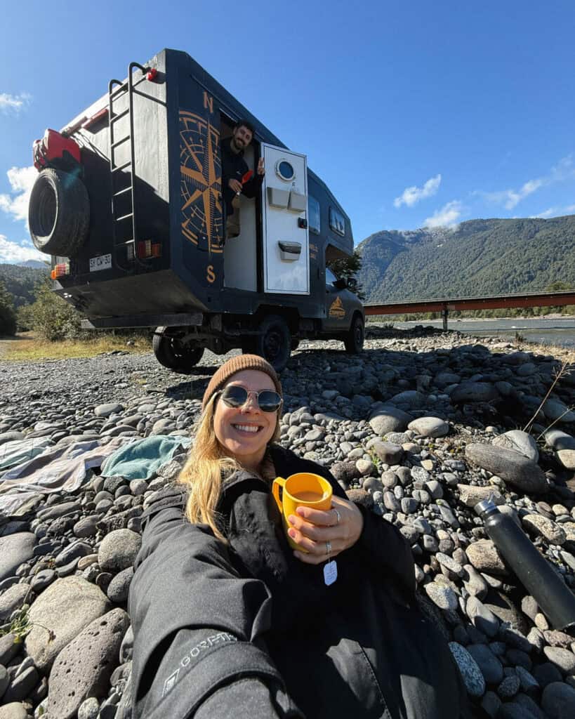 Chaiten, Chile Travel Guide: A Town That Nearly Disappeared 10 Sally smiles with a yellow cup while sitting on river rocks beside a camper truck in Chaitén, as Brayden leans out of the open door of the truck with forested mountains in the background.