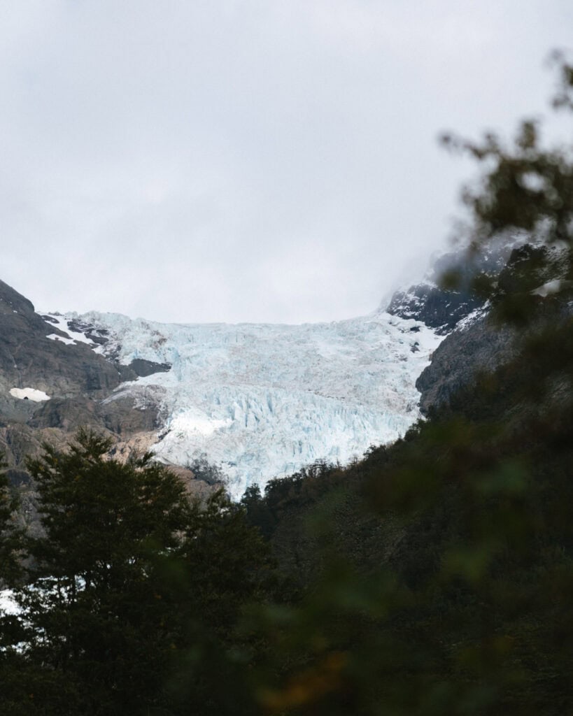 Ventisquero Yelcho Hike Guide: Corcovado National Park, Chile 1 Distant glacier in Chaitén peeking through a frame of forest trees, glowing pale blue against the rugged mountain backdrop.