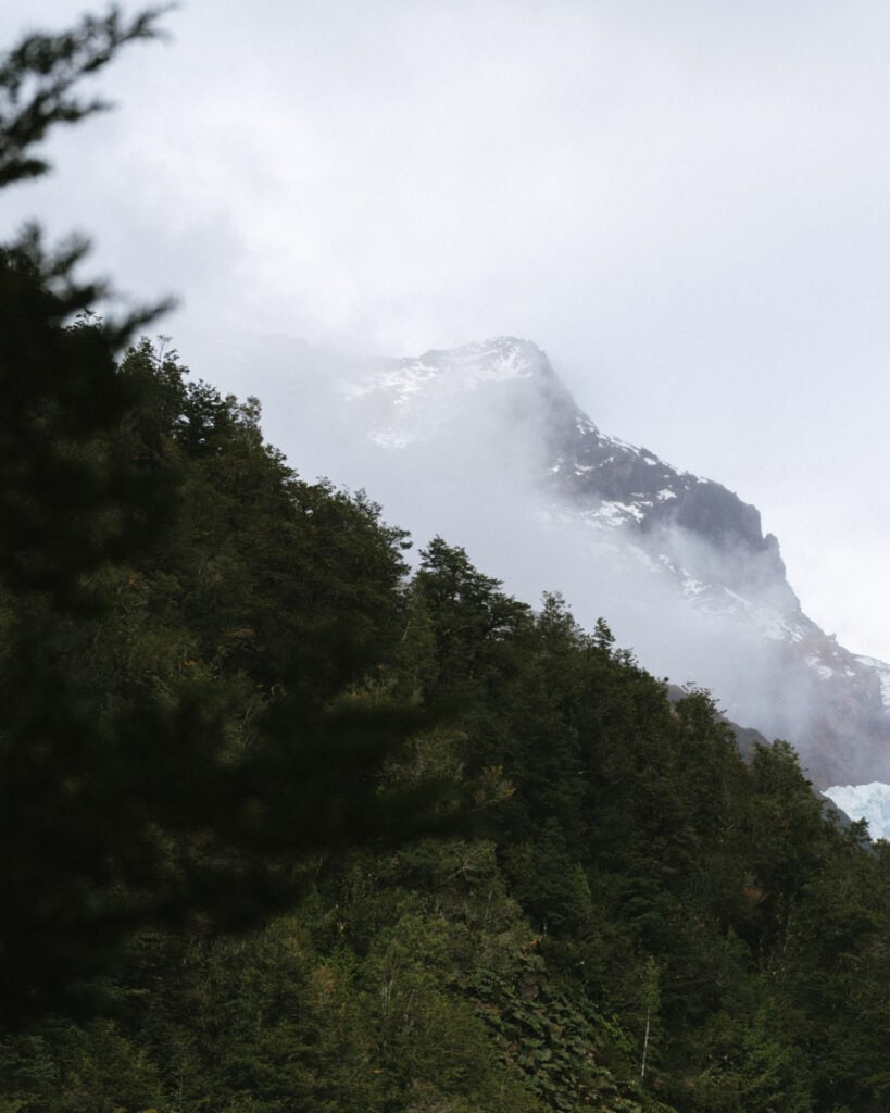 Ventisquero Yelcho Hike Guide: Corcovado National Park, Chile 10 Snow-dusted mountain peak in Chaitén partially hidden by swirling clouds and surrounded by dark green forest.