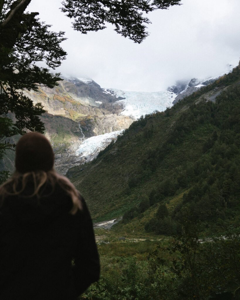 Ventisquero Yelcho Hike Guide: Corcovado National Park, Chile 20 Sally stands at the edge of a forest in Chaitén, Argentina, gazing toward the Ventisquero Yelcho glacier nestled between misty mountain peaks.