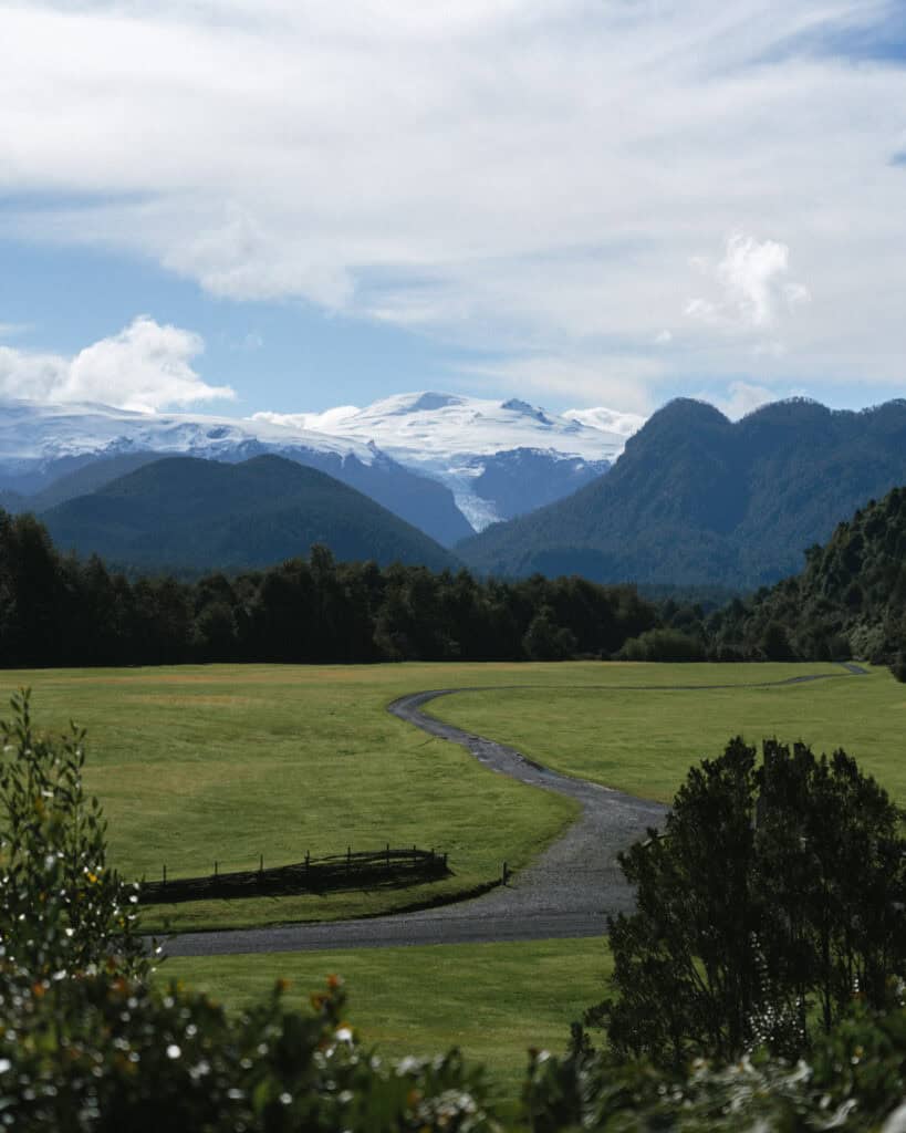 How to Visit the Pumalin National Park in Patagonia, Chile 43 Expansive grassy field in Chaitén with a winding path leading toward snow-capped mountains under a bright blue sky.