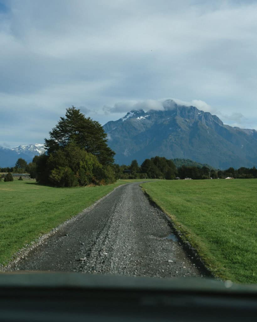 Chaiten, Chile Travel Guide: A Town That Nearly Disappeared 33 Gravel road stretches toward a towering snow-dusted mountain partly covered by clouds, flanked by green fields.