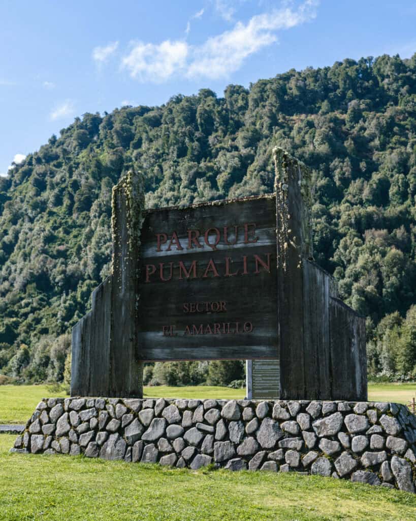How to Visit the Pumalin National Park in Patagonia, Chile 35 Large wooden sign for Parque Pumalín, Sector El Amarillo, mounted on a stone base in a grassy field with forested hills behind.