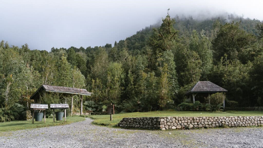 How to Visit the Pumalin National Park in Patagonia, Chile 18 Park entrance area with rustic shelters, stone-bordered paths, and trash bins surrounded by dense forest and mist-covered hills.