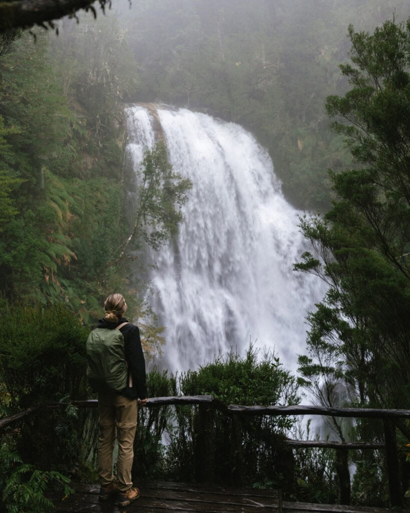 Chaiten, Chile Travel Guide: A Town That Nearly Disappeared 3 Sally in hiking gear with a green backpack stands on a wet wooden platform gazing at a large cascading waterfall.