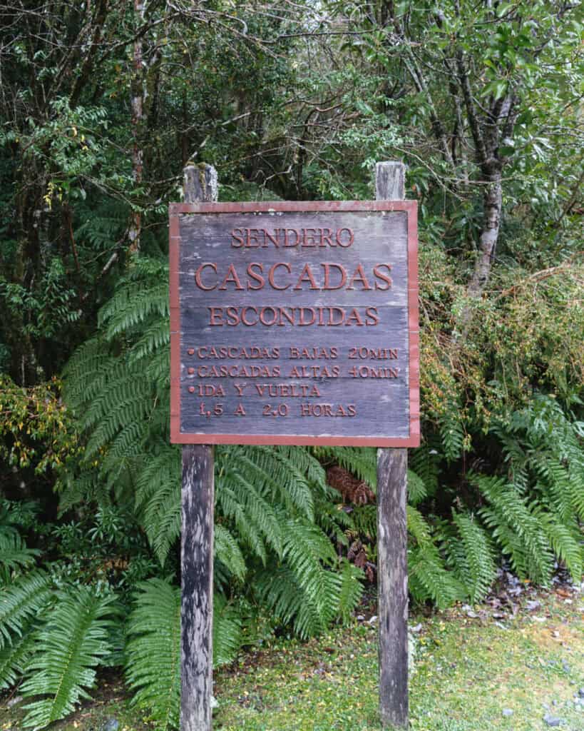How to Visit the Pumalin National Park in Patagonia, Chile 24 Wooden trailhead sign for Sendero Cascadas Escondidas with walking times listed for lower and upper waterfalls, surrounded by ferns and trees.
