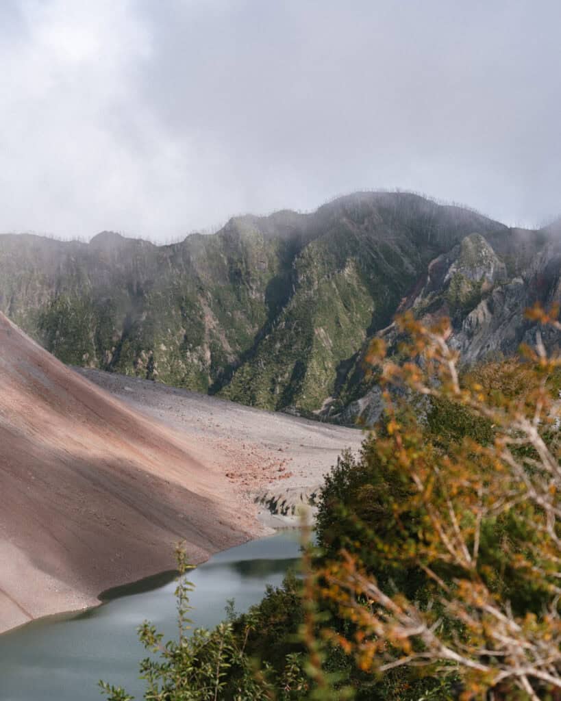 How to Visit the Pumalin National Park in Patagonia, Chile 2 Rust-red slope of the Chaitén Volcano meets a pale green lagoon under cloudy skies, showing volcanic rock and ash.