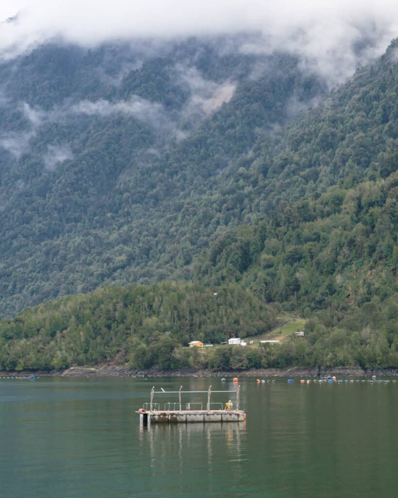Hornopiren, Chile Travel Guide: Termas, Alerces & Fjords 26 Floating boat in the calm waters of Hornopirén, Chile, with forested hills rising steeply in the background, partly covered by low-hanging mist and clouds.