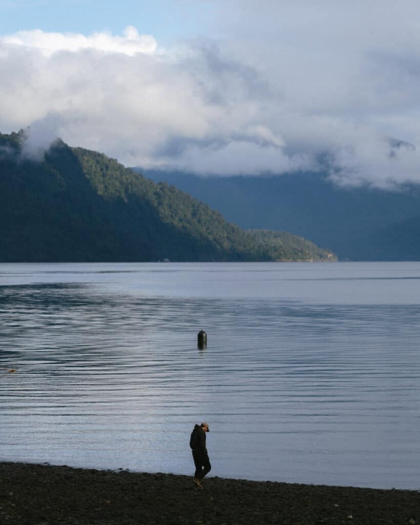 Hornopiren, Chile Travel Guide: Termas, Alerces & Fjords 9 Brayden walking along the rocky shore of Hornopirén, Chile, with still waters reflecting the surrounding misty green mountains under a cloudy sky.
