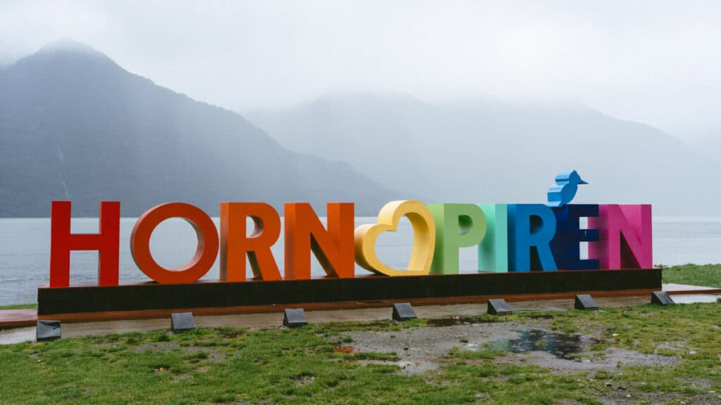 Hornopiren, Chile Travel Guide: Termas, Alerces & Fjords 24 Colorful rainbow sign reading “Hornopirén” with a bird symbol above the letters, set against a misty fjord and mountains in Hornopirén, Chile.