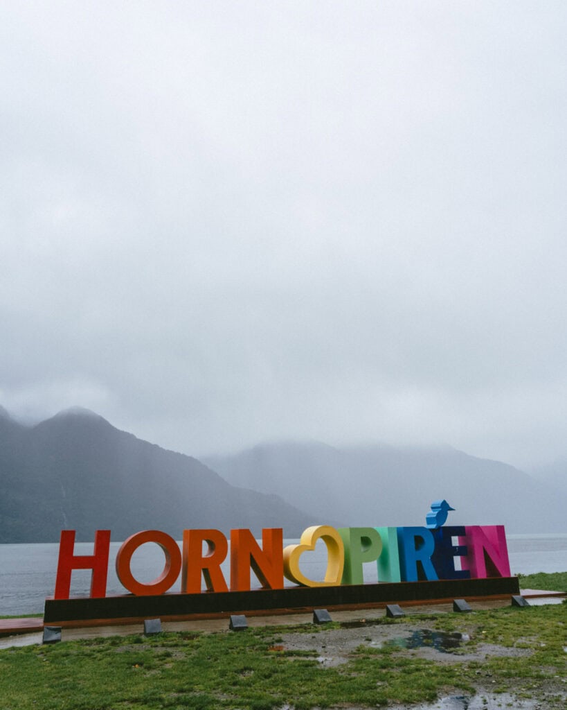 Hornopiren, Chile Travel Guide: Termas, Alerces & Fjords 4 Wide view of the rainbow-colored “Hornopirén” sign with mountains and water behind it, on a misty day in Hornopirén, Chile.