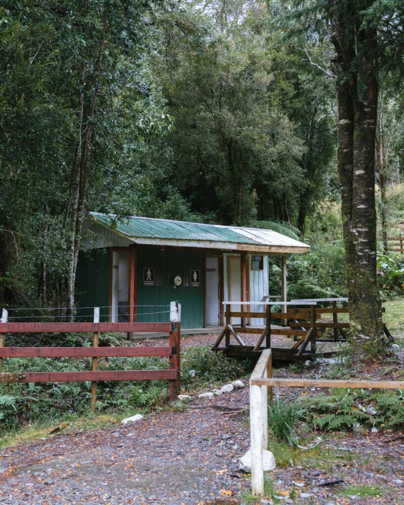 How to Visit the Alerce Andino National Park, Chile 10 Small green building with restrooms marked “Damas” and “Hombres,” located in a forested area of Hornopirén, Chile.
