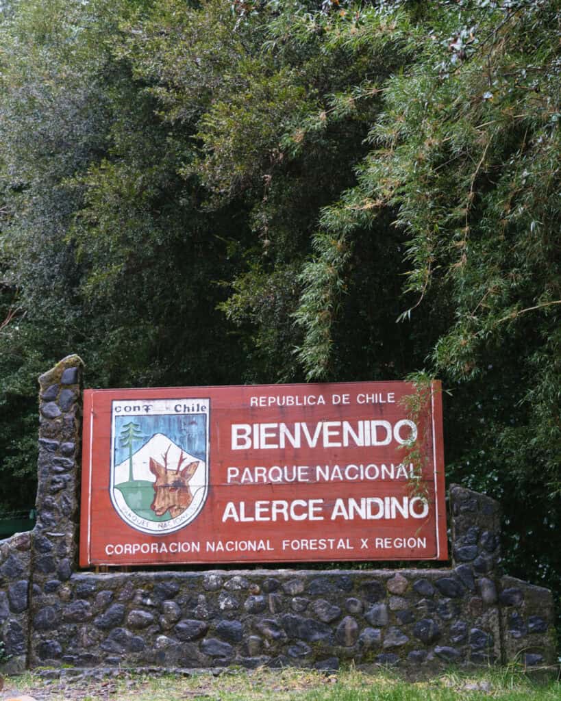 Hornopiren, Chile Travel Guide: Termas, Alerces & Fjords 29 Alternate view of the wooden welcome sign for Parque Nacional Alerce Andino, Chile, surrounded by lush green foliage.