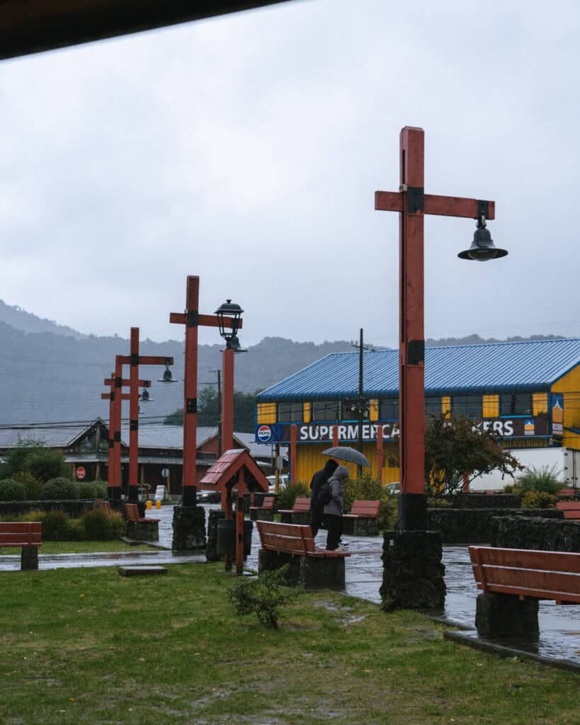 Hornopiren, Chile Travel Guide: Termas, Alerces & Fjords 33 Strangers walking under an umbrella in a plaza in Hornopirén, Chile, with red lampposts and benches lining the square.