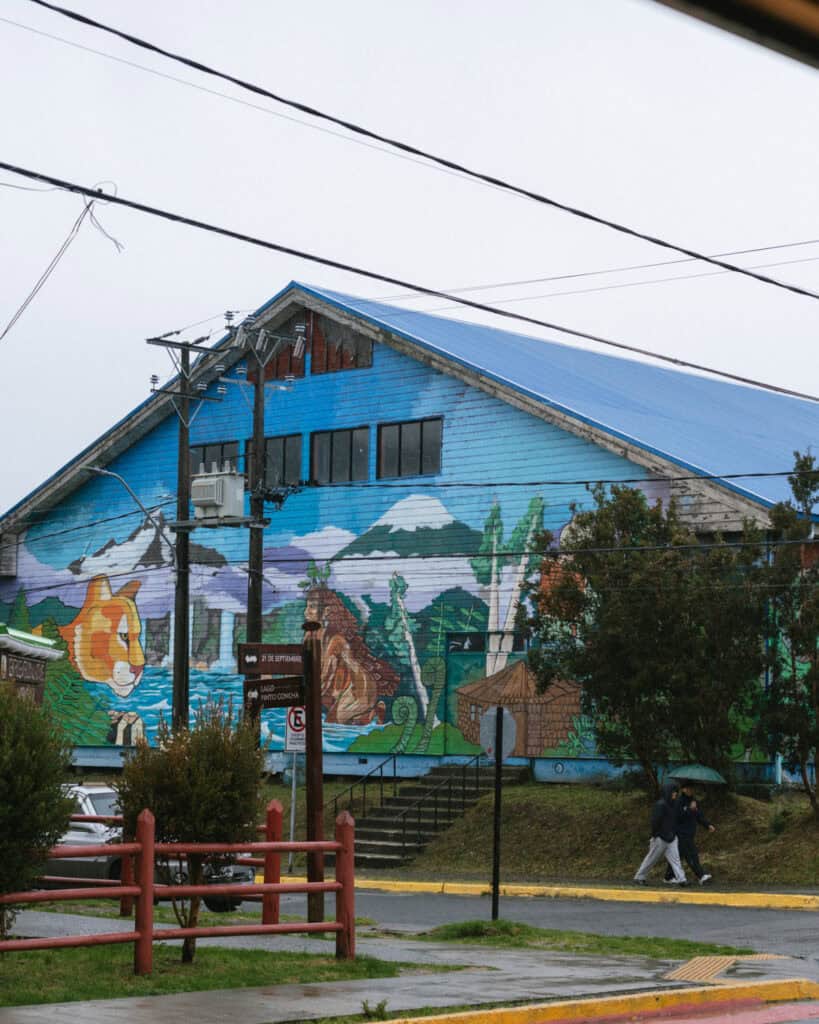 Hornopiren, Chile Travel Guide: Termas, Alerces & Fjords 23 Large mural painted on the side of a building in Hornopirén, Chile, showing a fox, landscapes, and cultural symbols with two people walking past under an umbrella.