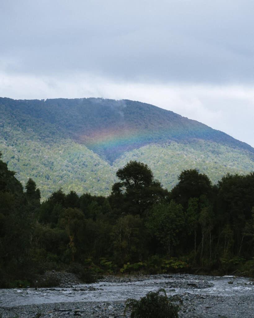 Hornopiren, Chile Travel Guide: Termas, Alerces & Fjords 13 Rainbow arching over a forested hillside with a rocky river in the foreground in Hornopirén, Chile.