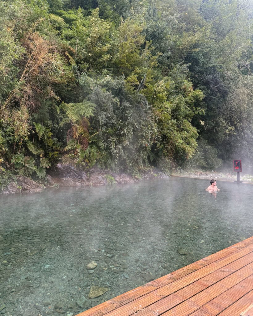 Puyuhuapi, Chile Travel Guide: Fjords and Hanging Glaciers 18 Wide hot spring pool with clear steaming water and Brayden bathing near the edge, set against dense forest in Hornopirén, Chile.