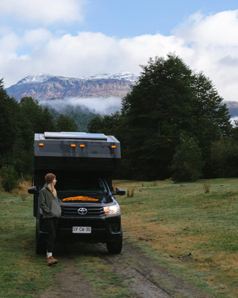 Puyuhuapi, Chile Travel Guide: Fjords and Hanging Glaciers 10 Sally standing beside a black Toyota camper truck parked on a grassy trail, with misty forests and snow-dusted mountains in the background in Puyuhuapi, Chile.
