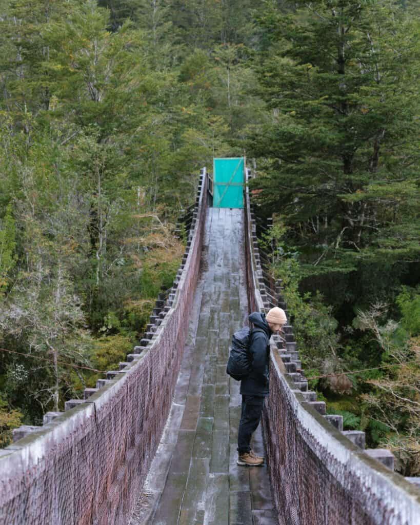 Puyuhuapi, Chile Travel Guide: Fjords and Hanging Glaciers 12 Hiker standing on a narrow wooden suspension bridge with red mesh sides, surrounded by dense forest in Parque Nacional Queulat, Chile.