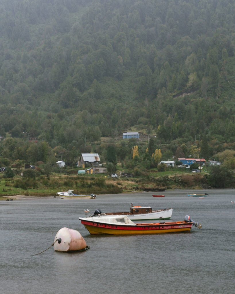 Puyuhuapi, Chile Travel Guide: Fjords and Hanging Glaciers 17 Small fishing boats and buoys floating near the shore of Puyuhuapi, Chile, with hillside houses and green forest in the distance.