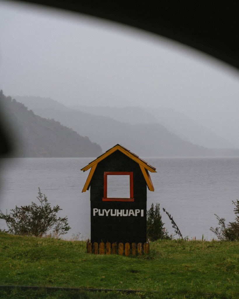 Puyuhuapi, Chile Travel Guide: Fjords and Hanging Glaciers 2 Wooden frame painted black with yellow roof and red trim displaying the word “Puyuhuapi” beside the fjord, serving as a town landmark.