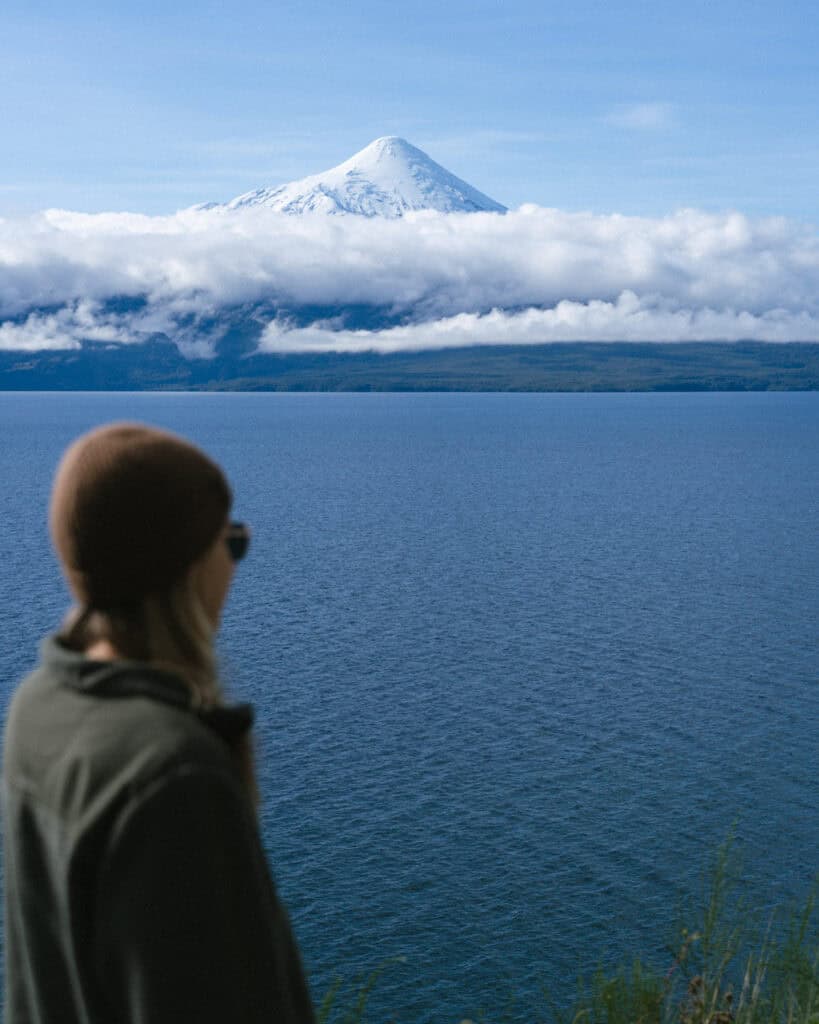 The Carretera Austral, Chile: Everything You Need to Know 21 Sally in a brown beanie and green jacket looking across the blue waters of Lake Llanquihue toward Osorno Volcano above the clouds.