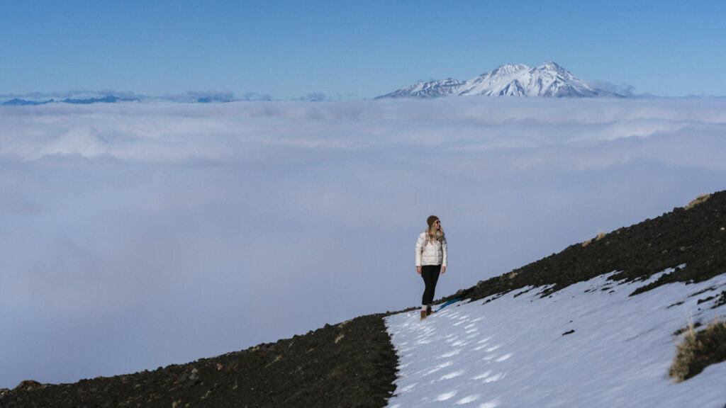 About Us 13 Sally walking along a narrow snow path on Osorno Volcano, with distant Andes mountains above the cloud line.