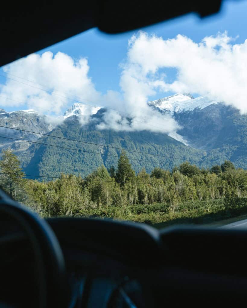 Chaiten, Chile Travel Guide: A Town That Nearly Disappeared 9 A shot of a glacier sitting on top of a forested mountain, seen through the front windscreen of a car, driving through Pumalin National Park. The steering wheel and dash are out of focus in the foreground.