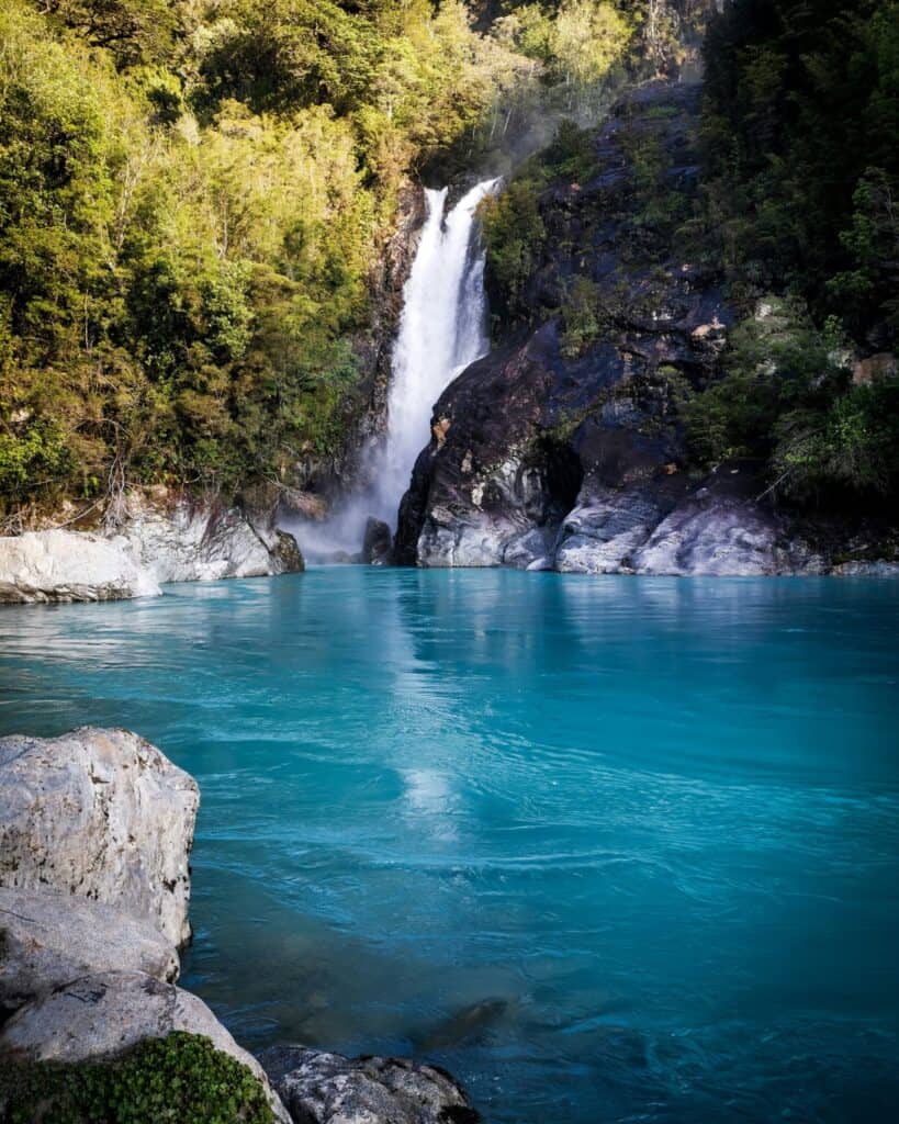Hornopiren, Chile Travel Guide: Termas, Alerces & Fjords 20 A narrow single drop waterfall falls amongst rocks into the vibrant blue Rio Blanco in Hornopiren National Park. The setting is surrounded by vibrant green forest.