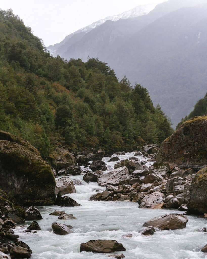 Queulat National Park: How to Visit the Ventisquero Colgante 4 The Rio Ventisquero in Parque Nacional Queulat, with rocks in the river, and a green forest on its slopes with a snow dusted mountain in the background.