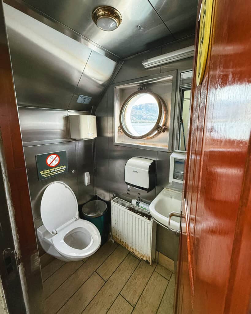 TABSA Ferry: Sailing from Puerto Natales to Puerto Yungay 26 Small metallic ferry bathroom on the Tabsa Ferry in Chile, with a toilet, compact sink, paper towel dispenser, and a round porthole window offering a view of the water and mountains.