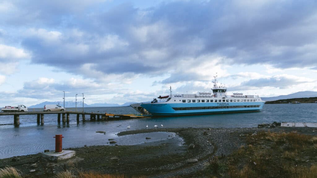 TABSA Ferry: Sailing from Puerto Natales to Puerto Yungay 5 Vehicles line up on a pier beside the Tabsa Ferry in Chile as it prepares for boarding, with a low tide revealing rocks and a few seabirds gathered near the shore.